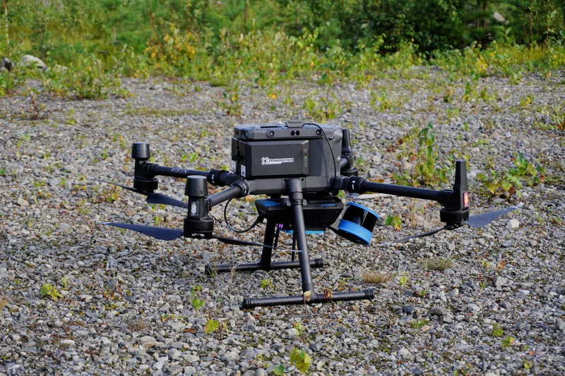 A black four-propeller drone fitted with a LiDAR sensor resting on gravel.
