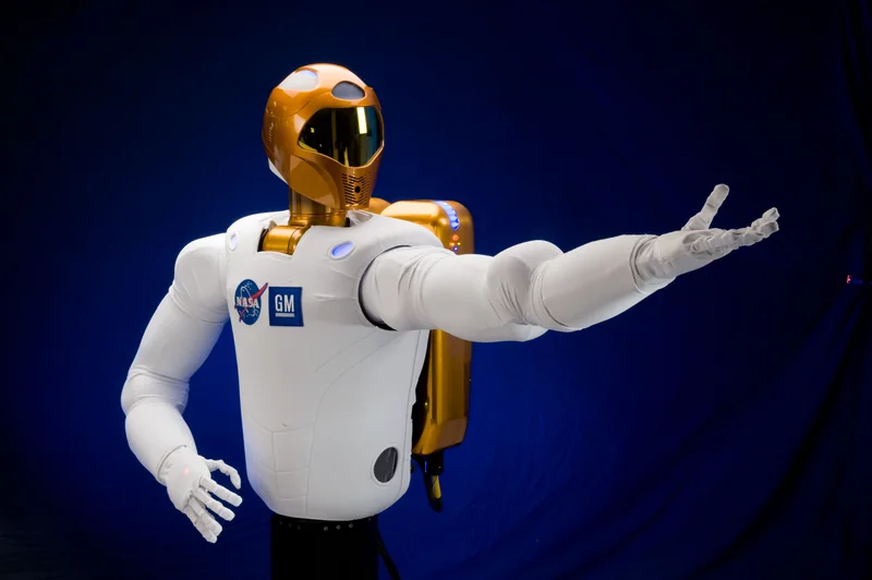 The white and gold Robonaut 2 humanoid research robot extending its left arm against a blue backdrop.